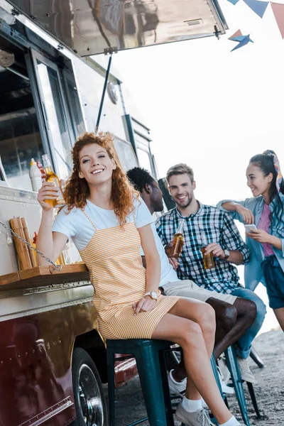 Счастливая рыжая девушка с бутылкой пива возле мультикультурных друзей и фургона с едой — стоковое фото