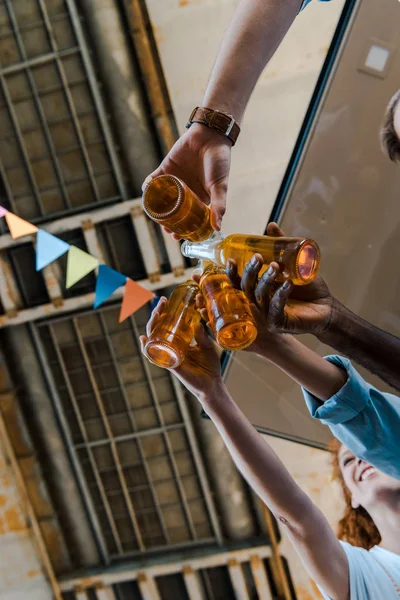 Vista inferior de amigos multiculturales felices tintinear botellas con cerveza - foto de stock