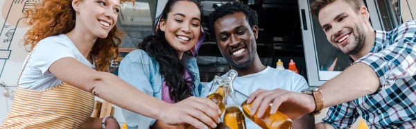 Panoramic shot of happy multicultural friends clinking bottles with beer near food truck — Stock Photo