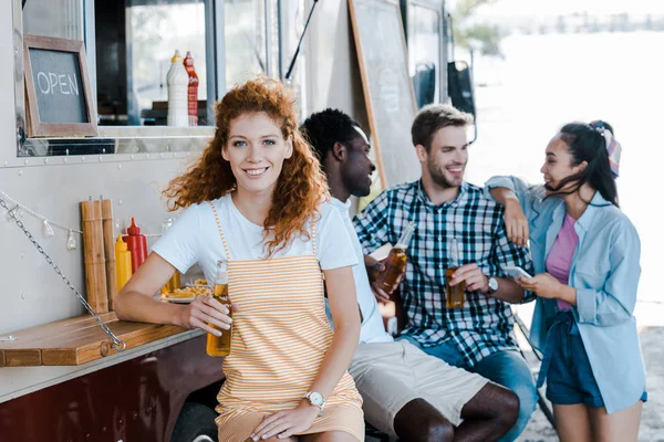 Enfoque selectivo de chica pelirroja feliz sosteniendo botella con cerveza cerca de amigos y camión de comida - foto de stock