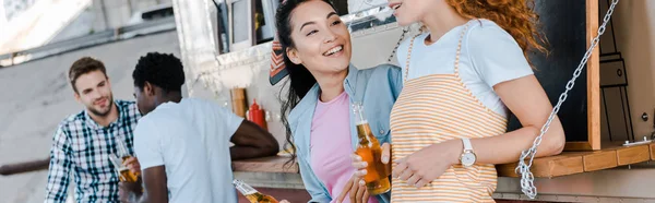 Panoramic shot of happy girls holding bottles with beer near multicultural men and food truck — Stock Photo