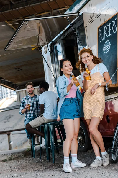 Mulheres felizes segurando garrafas com cerveja enquanto está perto de homens multiculturais e caminhão de alimentos — Fotografia de Stock