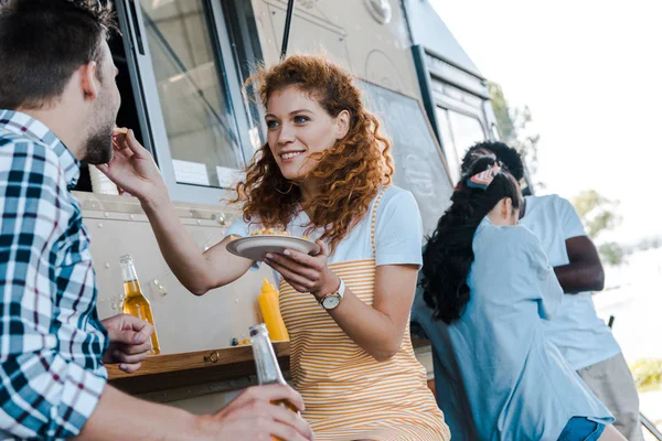 Selective focus of redhead girl feeding man near multicultural people — Stock Photo