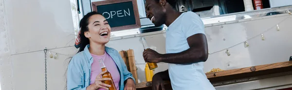 Panoramic shot of asian girl laughing near african american man with bottle of beer — Stock Photo