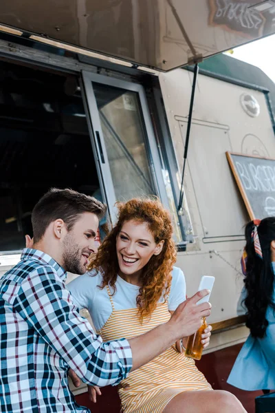 Selective focus of redhead girl looking at man holding smartphone near food truck — Stock Photo