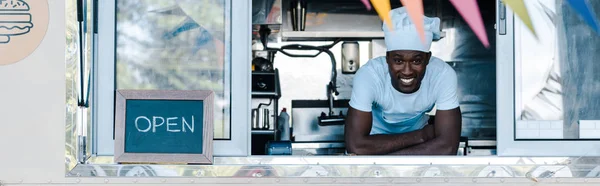 Colpo panoramico di felice uomo africano americano in uniforme da chef sorridente da camion cibo — Foto stock