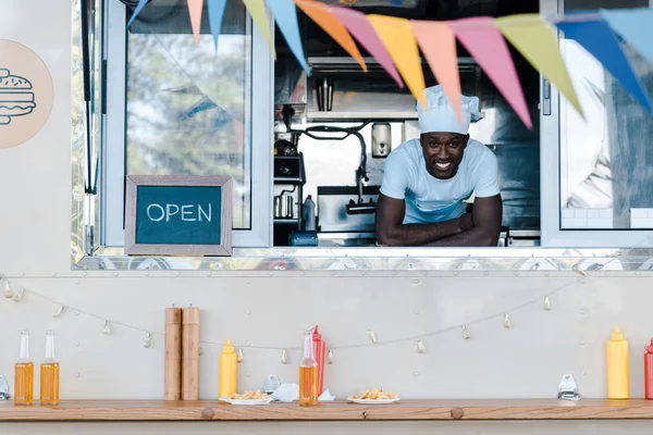 Alegre africano americano hombre en chef uniforme sonriendo desde comida camión - foto de stock