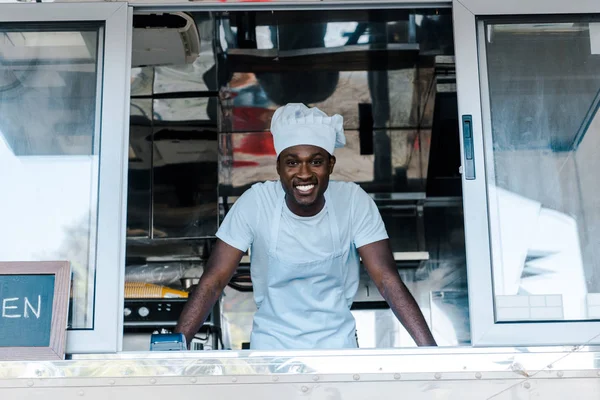 Homme afro-américain joyeux en uniforme de chef et chapeau souriant de camion alimentaire — Photo de stock