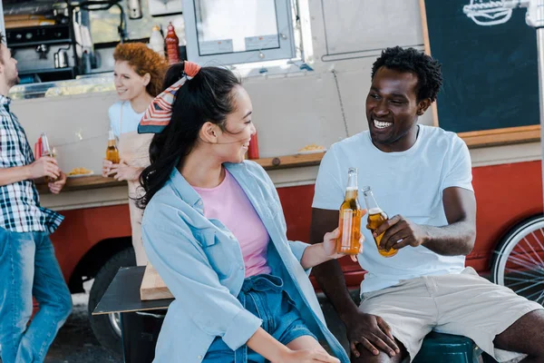 Selective focus of cheerful asian girl smiling near african american man and clinking bottles of beer near food truck and people — Stock Photo