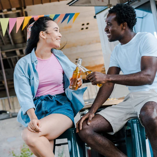 Веселая азиатская женщина улыбается рядом с африканским американским мужчиной и звенящие бутылки пива возле фургона с едой — стоковое фото