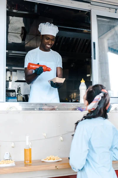 Селективное внимание африканского американца, держащего картонную тарелку и бутылку кетчупа рядом с клиентом — стоковое фото