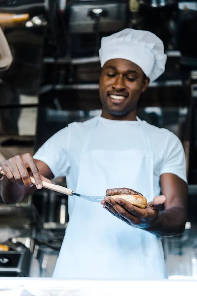 Selective focus of handsome afrian american man holding spatula while preparing burger — Stock Photo