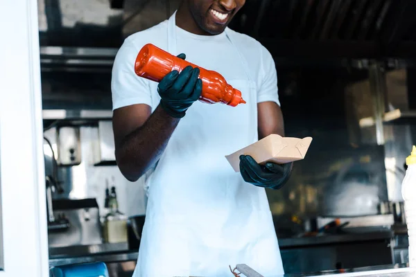 Vista recortada de hombre americano africano alegre sosteniendo botella de ketchup cerca de plato - foto de stock