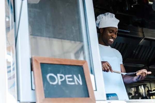 Selective focus of happy afrian american man holding spatula while preparing burger near chalk board with open letters — Stock Photo