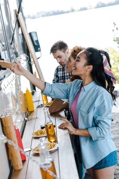 Cropped view of african american man giving carton plate to asian girl near food truck and people — Stock Photo