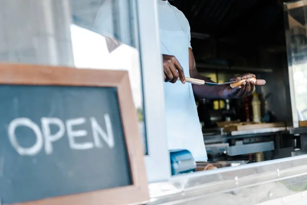 Vista recortada del hombre afriano americano sosteniendo espátula mientras prepara hamburguesa cerca de pizarra con letras abiertas - foto de stock