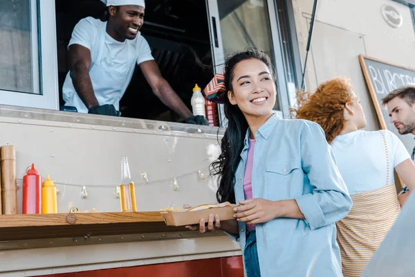 Enfoque selectivo de alegre chica asiática sosteniendo placa de cartón cerca de clientes y chef en camión de comida - foto de stock