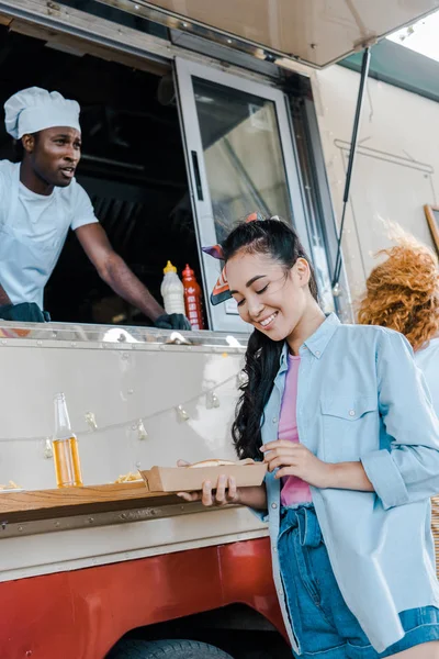 Enfoque selectivo de alegre chica asiática mirando cartón plato cerca de chef en camión de comida - foto de stock