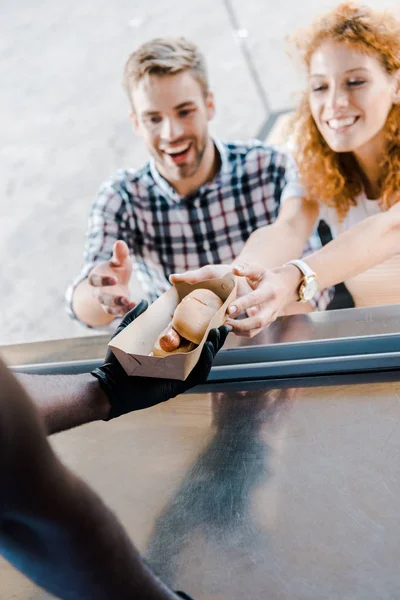 Vue recadrée de l'homme afro-américain donnant plaque de carton avec hot dog aux clients heureux — Photo de stock