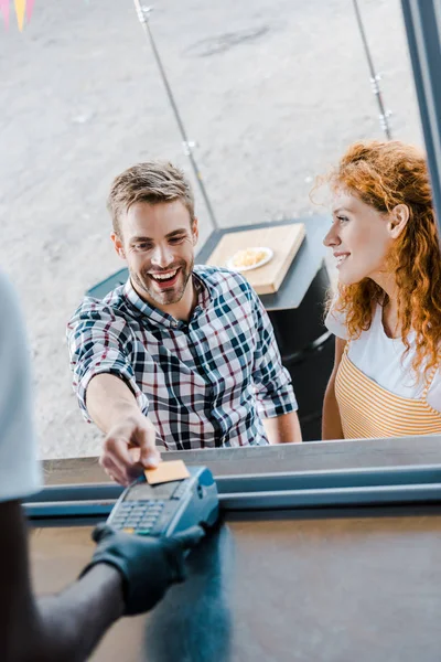 Vista recortada del hombre afroamericano sosteniendo lector de tarjetas de crédito cerca de clientes con tarjeta de crédito - foto de stock