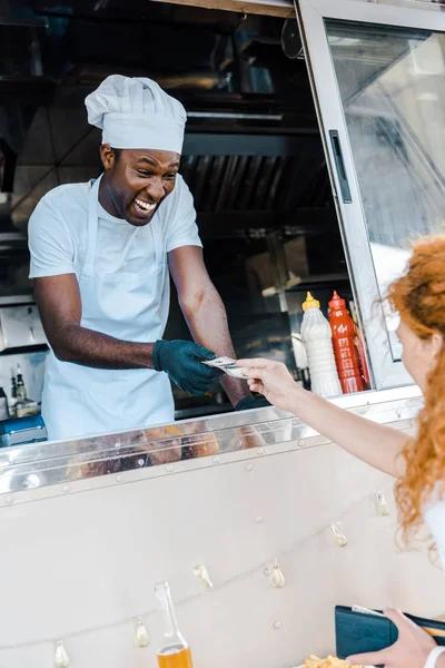 Selettiva messa a fuoco di rossa ragazza dando denaro contante per felice africano americano uomo in cibo camion — Foto stock