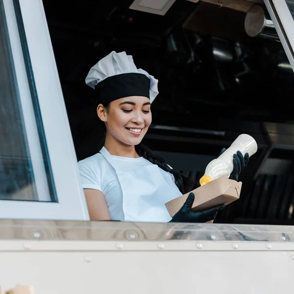 Low angle view of happy asian woman holding carton plate and mayonnaise bottle in food truck — Stock Photo