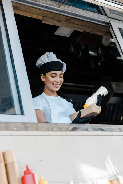 Low angle view of cheerful asian woman holding carton plate and mayonnaise bottle in food truck — Stock Photo