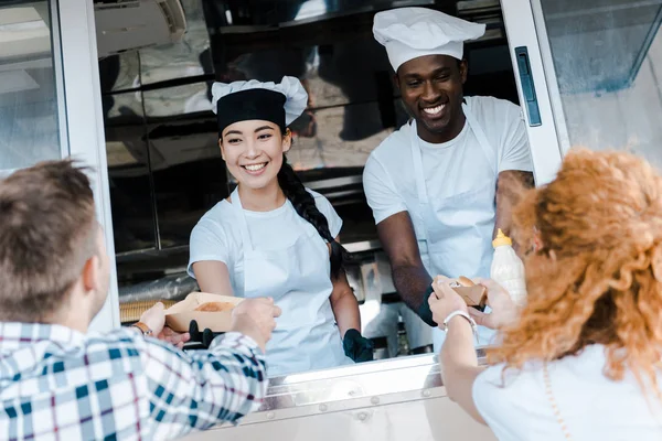 Enfoque selectivo de chefs multiculturales felices dando platos de cartón con alimentos a los clientes - foto de stock