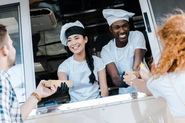 Enfoque selectivo de chefs multiculturales alegres que dan platos de cartón con alimentos a los clientes - foto de stock