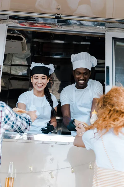 Enfoque selectivo de chefs multiculturales sonrientes dando platos de cartón con comida a los clientes - foto de stock