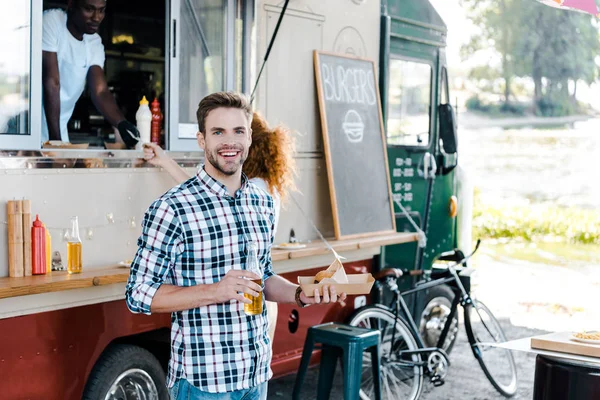 Selective focus of happy man holding bottle with beer and carton plate with street food near woman and african american man in food truck — Stock Photo