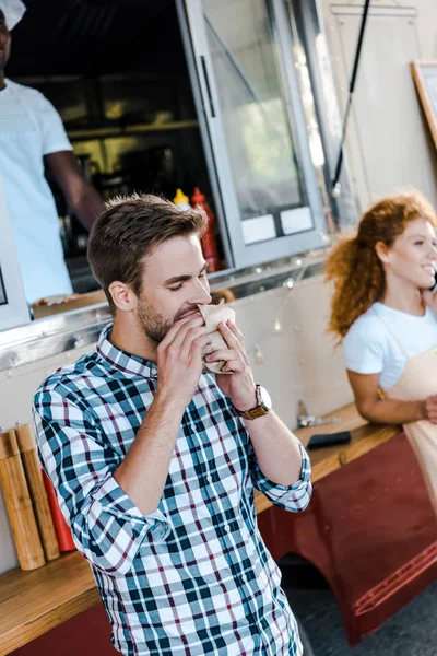 Selektiver Fokus eines gutaussehenden Mannes, der in der Nähe eines Foodtrucks Burger isst — Stockfoto