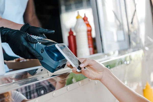 Vista recortada de la mujer que paga con teléfono inteligente cerca de chica en camión de comida - foto de stock