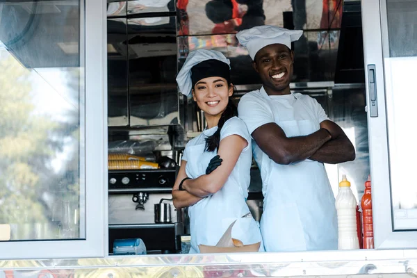 Asian girl and african american man in hats smiling while standing with crossed arms in food truck — Stock Photo