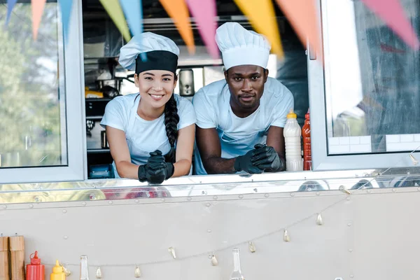 Selective focus of asian girl and african american man in hats smiling from food truck — Stock Photo