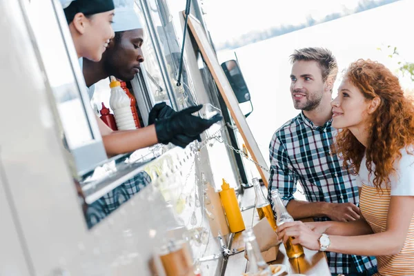 Enfoque selectivo de los chefs multiculturales mirando al hombre y la mujer con cerveza - foto de stock