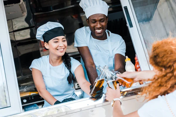 Enfoque selectivo de los chefs multiculturales tintineo con botellas de cerveza cerca de los clientes - foto de stock