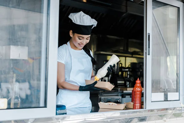 Cheerful asian woman holding carton plate and mayonnaise bottle in food truck — Stock Photo