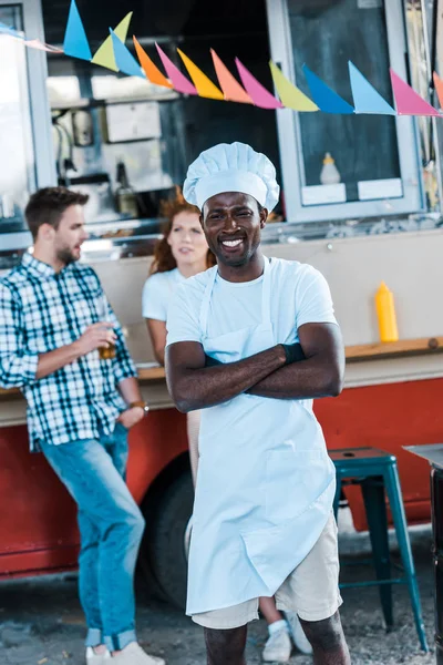 Foco seletivo do chef americano africano feliz de pé com braços cruzados perto de caminhão de alimentos e clientes — Fotografia de Stock