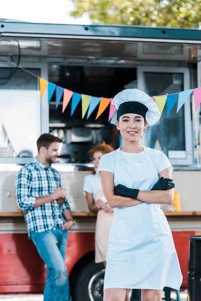 Selettivo fuoco di felice asiatico chef in piedi con braccia incrociate vicino cibo camion e clienti — Foto stock