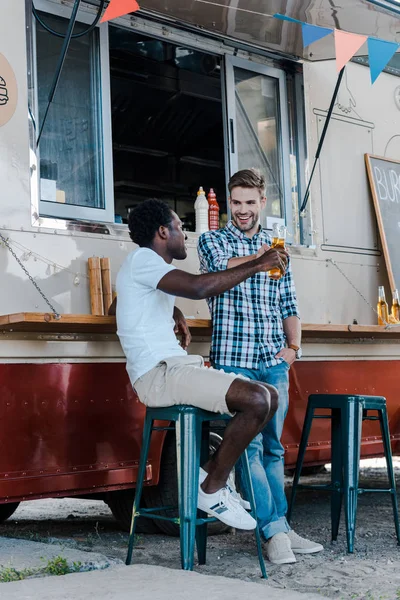 Happy multicultural men clinking bottles with beer near food truck — Stock Photo