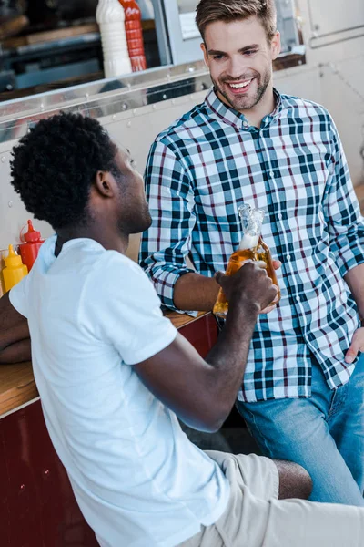 Selective focus of happy multicultural men clinking bottles with beer — Stock Photo