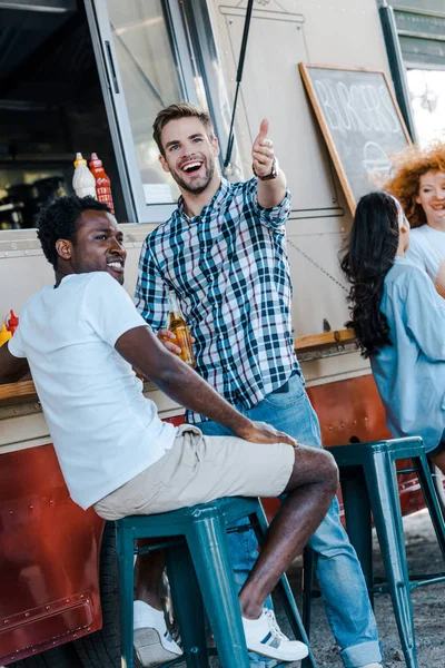 Foco seletivo do homem feliz gesticulando perto de amigo afro-americano e caminhão de comida — Fotografia de Stock