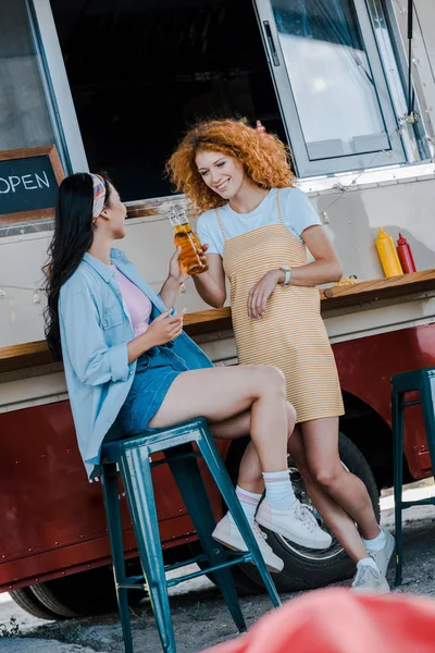 Happy multicultural girls holding bottles with beer near food truck — Stock Photo