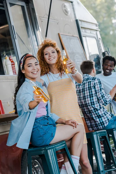 Selective focus of happy multicultural girls holding bottles of beer near food truck — Stock Photo