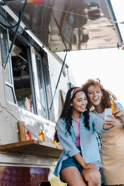 Foco seletivo de meninas multiculturais alegres tomando selfie perto de caminhão de alimentos — Fotografia de Stock