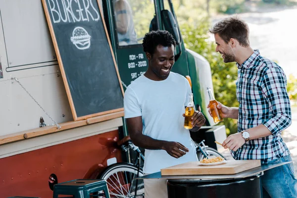 Happy multicultural men holding bottles of beer near food truck outside — Stock Photo