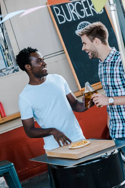 Positive multicultural men toasting bottles of beer near french fries and food truck — Stock Photo