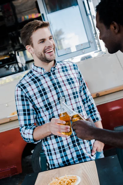 Selective focus of cheerful multicultural men toasting bottles of beer near french fries and food truck — Stock Photo