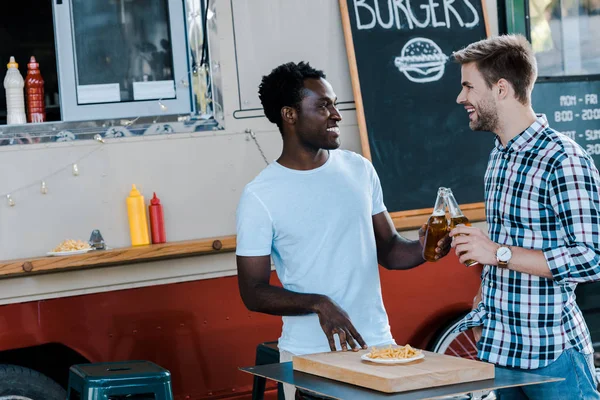 Sorrindo amigos multiculturais brindar garrafas de cerveja perto de batatas fritas e caminhão de alimentos — Fotografia de Stock